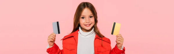 Chica sonriente en chaqueta naranja posando con tarjetas de crédito aisladas en rosa, pancarta - foto de stock