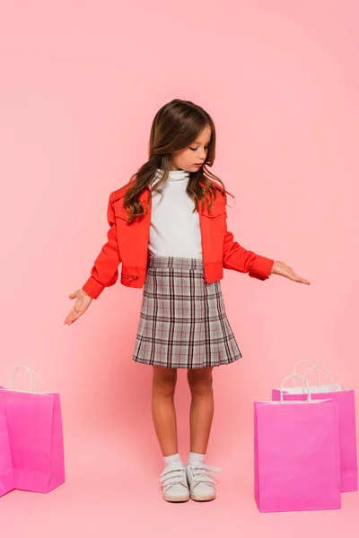 Mädchen in orangefarbener Jacke und kariertem Rock zeigt auf Einkaufstüten in rosa — Stockfoto