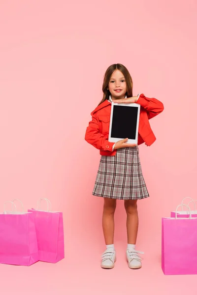 Enfant à la mode debout près des sacs à provisions avec tablette numérique sur rose — Photo de stock