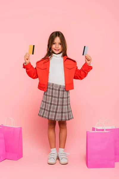 Стильна і щаслива дитина, що показує кредитні картки, стоячи поруч з сумками для покупок на рожевому — стокове фото