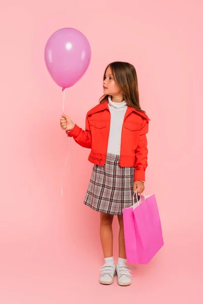 Fille en veste orange et jupe à carreaux debout avec ballon et sac à provisions sur rose — Photo de stock