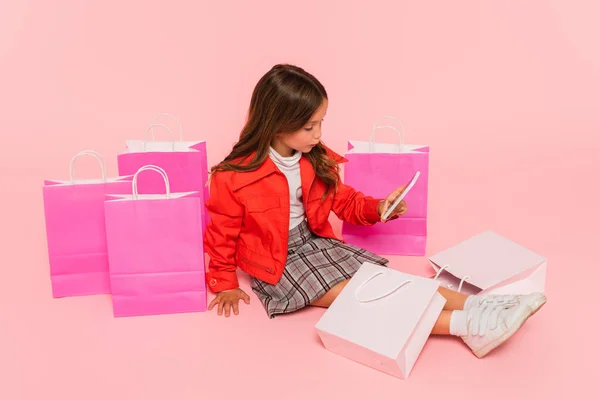 Fille en vêtements à la mode assis près de sacs à provisions sur rose — Photo de stock