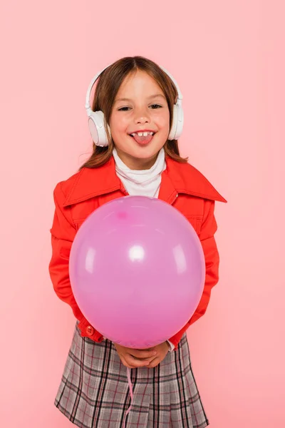 Alegre chica en auriculares sobresaliendo lengua mientras sostiene globo aislado en rosa - foto de stock