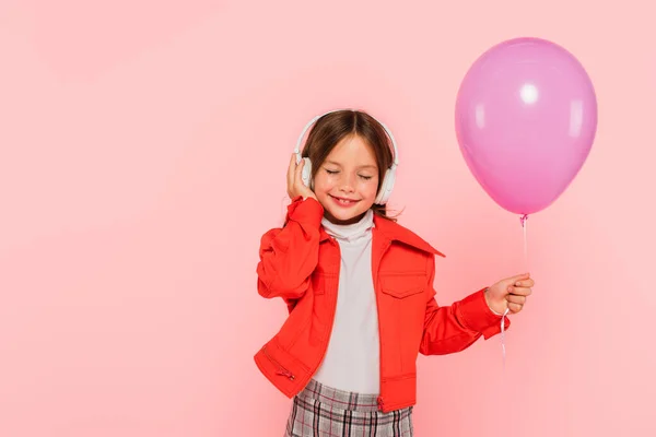 Chica feliz escuchando música con los ojos cerrados mientras sostiene globo festivo aislado en rosa - foto de stock