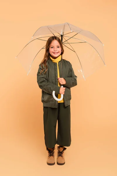 Повний вигляд усміхненої дівчини в бомбардувальнику, що стоїть під парасолькою на бежевому — стокове фото