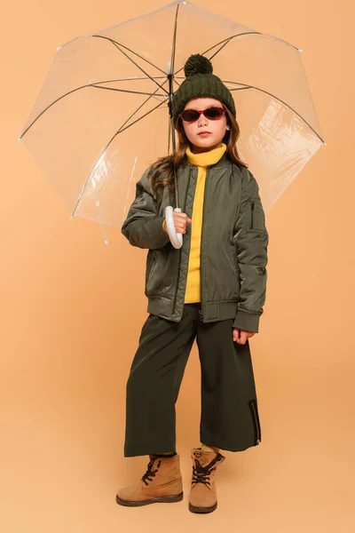 Vue pleine longueur de fille en veste de bombardier et bottes en daim sous parapluie transparent sur beige — Photo de stock