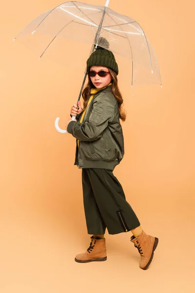 Child in fashionable autumn clothes posing under transparent umbrella on beige — Stock Photo