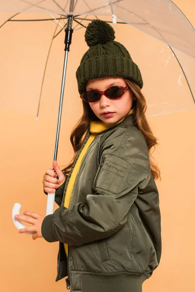 Kid in sunglasses and trendy autumn clothes under transparent umbrella isolated on beige — Stock Photo