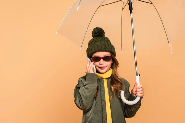 Niño con estilo en gafas de sol hablando en el teléfono inteligente bajo paraguas aislado en beige - foto de stock