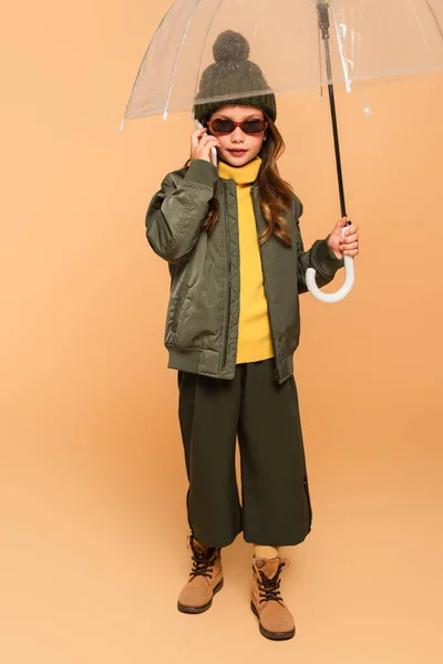 Full length view of girl in trendy clothes talking on cellphone under umbrella on beige — Stock Photo