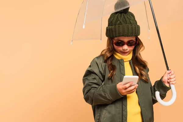 Menina em roupa de outono na moda e óculos de sol usando smartphone sob guarda-chuva isolado no bege — Fotografia de Stock