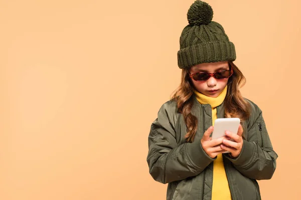 Girl in trendy autumn outfit and sunglasses using smartphone isolated on beige — Stock Photo