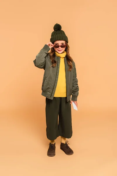 Cheerful girl in fashionable autumn clothes taking off sunglasses while looking at camera on beige — Stock Photo