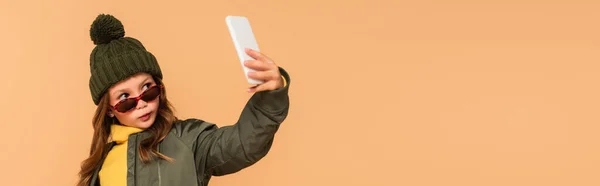 Menina na moda em chapéu de malha e óculos de sol tomando selfie isolado em bege, banner — Fotografia de Stock