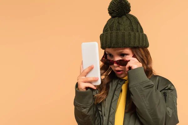 Chica sorprendida quitándose gafas de sol con estilo mientras mira el teléfono celular aislado en beige - foto de stock
