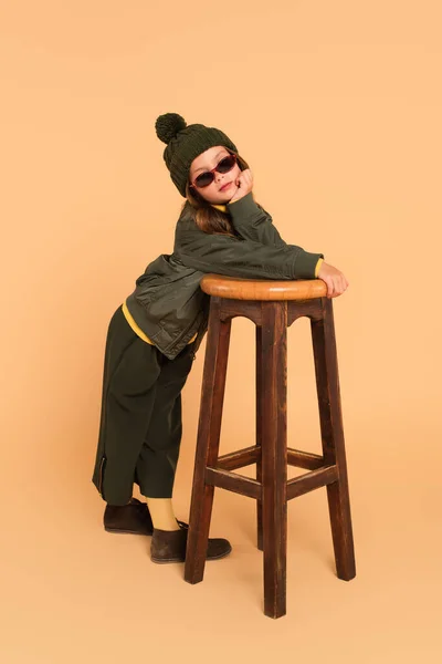 Kid in sunglasses, bomber jacket, pants and knitted hat posing near wooden stool on beige — Stock Photo