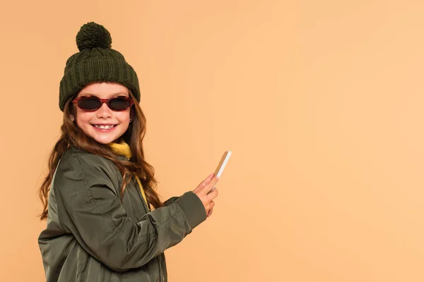 Cheerful kid in sunglasses, jacket and knitted hat holding mobile phone isolated on beige — Stock Photo