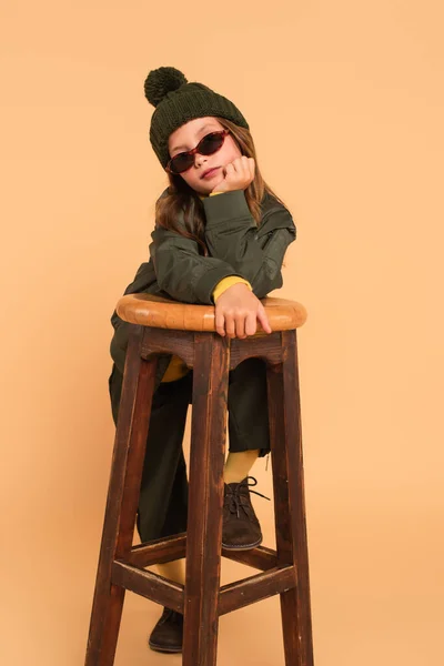 Criança em óculos de sol e roupas de outono na moda olhando para a câmera perto de banquinho de madeira no bege — Fotografia de Stock