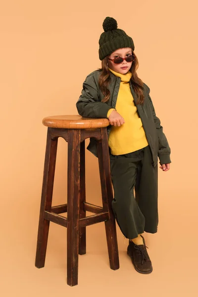 Child in yellow turtleneck and bomber jacket posing near high stool on beige — Stock Photo