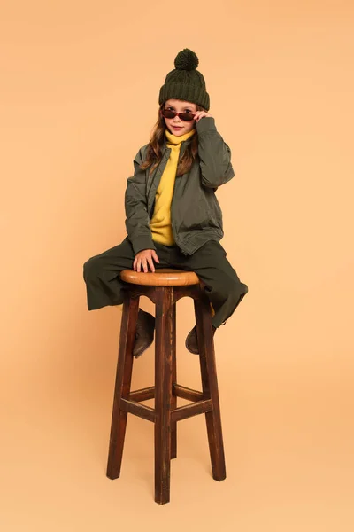 Trendiges Kind blickt über Sonnenbrille in die Kamera, während es auf einem hohen Holzhocker in Beige sitzt — Stockfoto