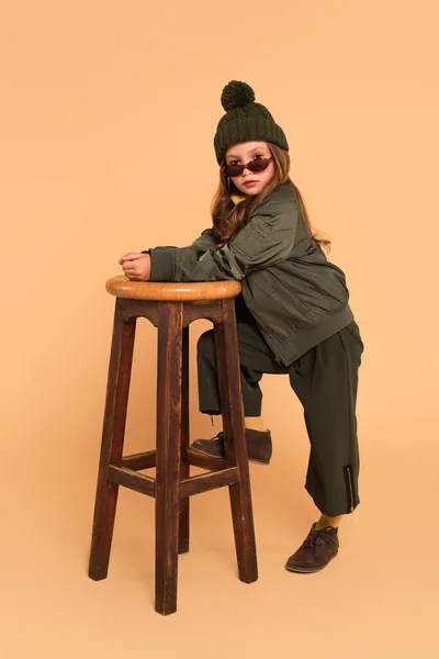 Fashionable girl in autumn clothes looking at camera over sunglasses near high stool on beige — Stock Photo