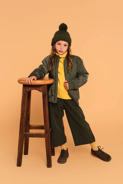 Stylish child in autumn outfit looking at camera near high wooden stool on beige — Stock Photo