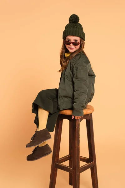 Усміхнена дитина в сонцезахисних окулярах і стильний осінній одяг, що сидить на високому табуреті на бежевому — стокове фото