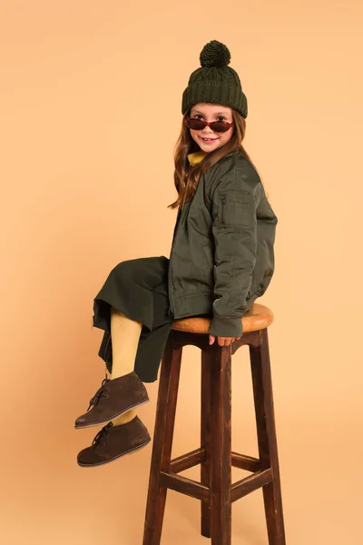 Smiling child in sunglasses and stylish autumn outfit sitting on high stool on beige — Stock Photo