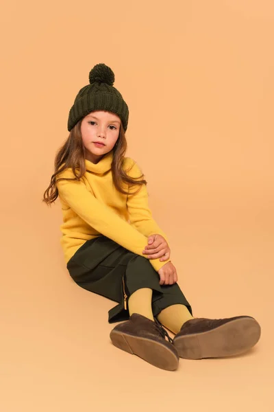 Trendy girl in yellow turtleneck and brown hat sitting on beige — Stock Photo
