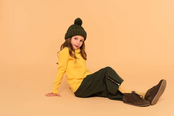 Vue pleine longueur de l'enfant en col roulé jaune, chapeau et pantalon tricotés assis sur beige — Photo de stock