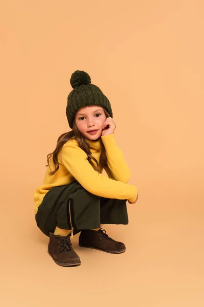 Ragazza in maglia cappello, collo alto giallo e stivali scamosciati guardando la fotocamera su beige — Foto stock