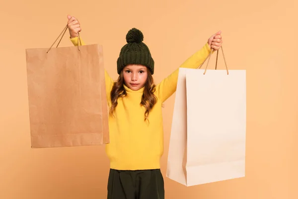 Chica de moda en cuello alto amarillo de pie con bolsas de compras en manos levantadas aisladas en beige - foto de stock