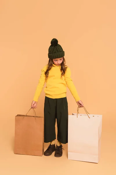 Полный вид на модного ребенка в желтой водолазке, стоящего с пакетами покупок на бежевом — стоковое фото