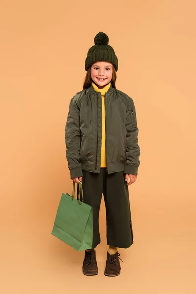 Happy girl in bomber jacket and brown hat holding shopping bag on beige — Stock Photo