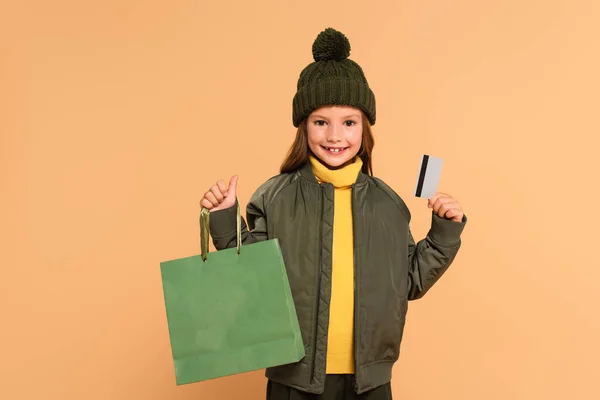 Allegra ragazza in bomber giacca e cappello a maglia che mostra carta di credito e shopping bag isolato su beige — Foto stock
