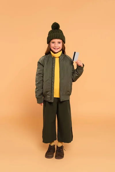 Visão de comprimento total da menina feliz em roupas de outono elegantes segurando cartão de crédito no bege — Fotografia de Stock