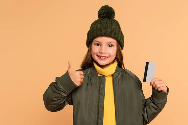 Niño feliz y elegante que muestra el pulgar hacia arriba mientras sostiene la tarjeta de crédito aislada en beige - foto de stock