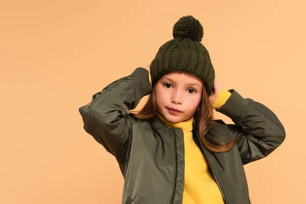 Niño en sombrero marrón y chaqueta posando con las manos detrás de la cabeza aisladas en beige - foto de stock