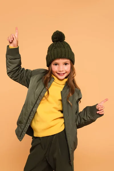 Bambino allegro in giacca e cappello a maglia che punta con le dita isolate sul beige — Foto stock