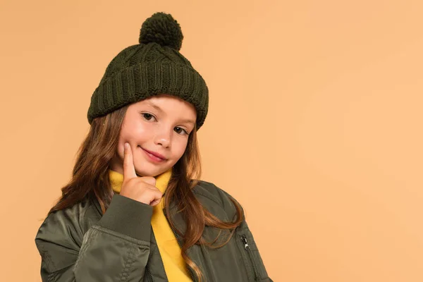 Capretto alla moda in cappello lavorato a maglia che tocca il viso mentre guarda la fotocamera isolata sul beige — Foto stock