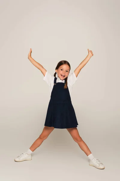 Cheerful schoolchild looking at camera while standing with raised hands on grey — Stock Photo