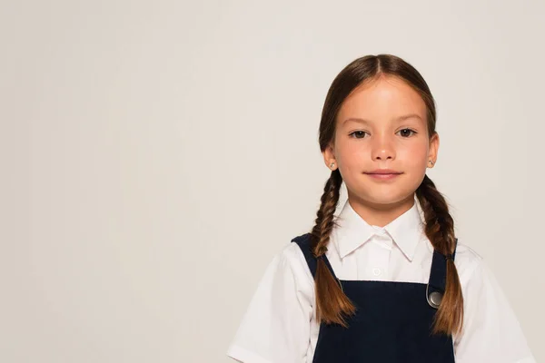 Позитивна дівчина в шкільній формі дивиться на камеру ізольовано на сірому — стокове фото