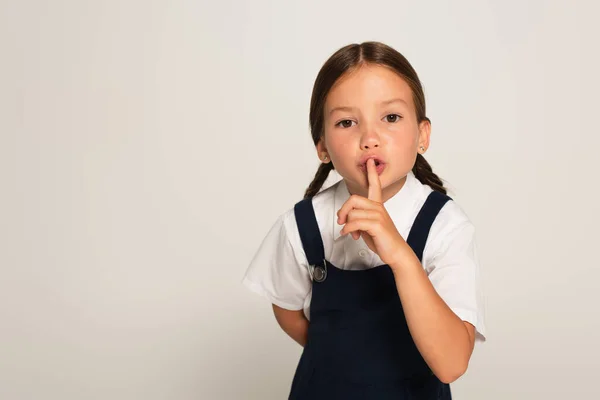 Girl in school uniform showing hush sign while looking at camera isolated on grey — Stock Photo