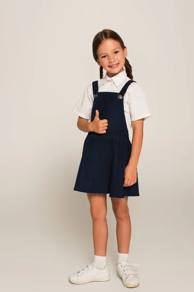 Alegre chica en uniforme escolar mostrando pulgar hacia arriba en gris - foto de stock