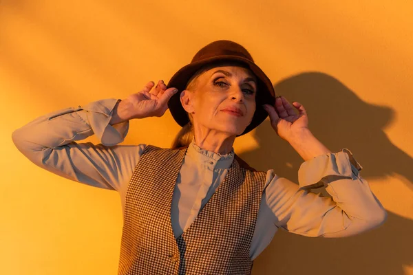 Femme âgée en gilet à carreaux tenant un chapeau sur fond orange — Photo de stock