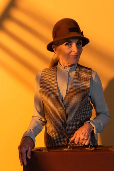 Donna anziana in abiti vintage con valigia su sfondo arancione — Foto stock