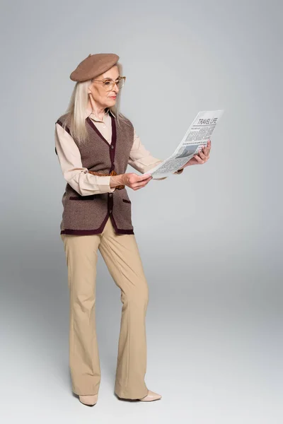 Stylish elderly woman reading travel newspaper on grey background — Stock Photo