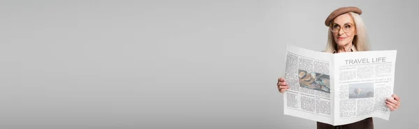 Elderly woman in beret and eyeglasses holding newspaper isolated on grey, banner — Stock Photo