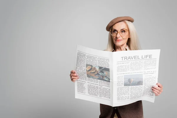 Elderly woman in beret holding newspaper with travel life lettering isolated on grey — Stock Photo