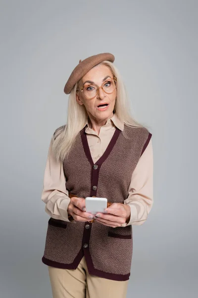 Amazed senior woman holding smartphone and looking at camera isolated on grey — Stock Photo